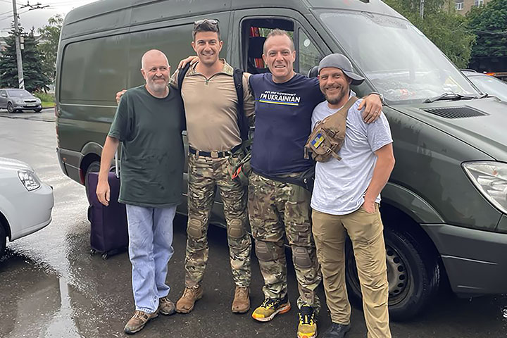 Brian Moscioni stands in front of a dark-colored van side-by-side with three of his fellow humanitarian volunteers
