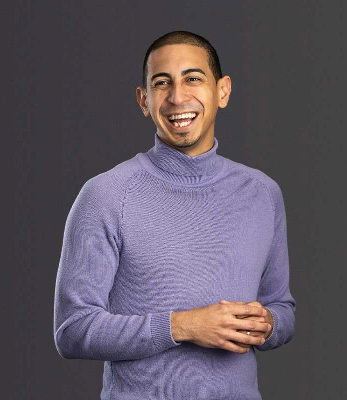 Associate Professor Alexander Diaz-Lopez stands smiling with his hands clasped, wearing a lavender turtleneck and black pants